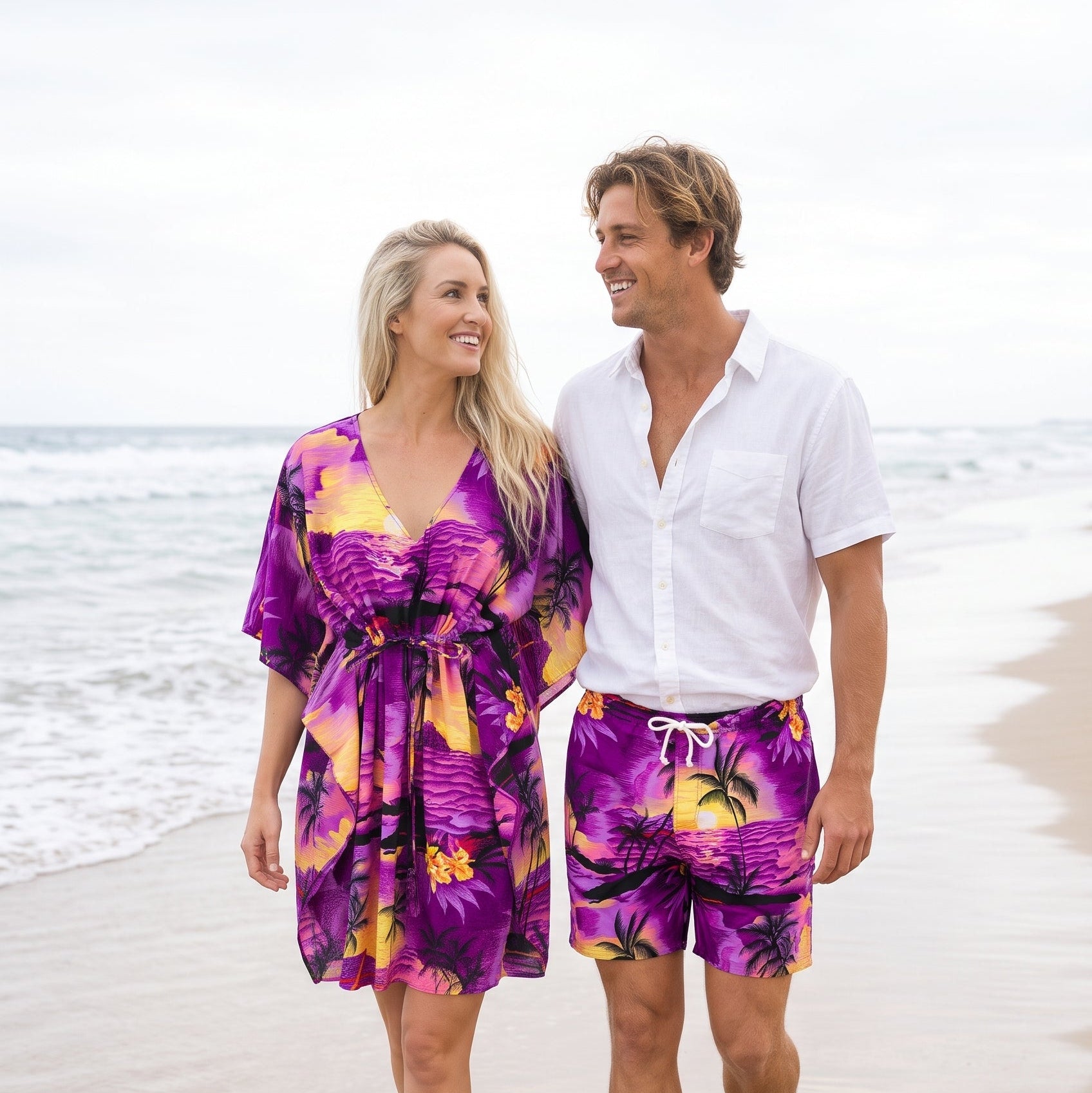 A couple walking on a sandy beach. The woman wears a purple tropical floral print kimono-style cover-up dress, and the man wears matching purple tropical print swim shorts with a white drawstring and a white button-down shirt.