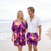 A couple walking on a sandy beach. The woman wears a purple tropical floral print kimono-style cover-up dress, and the man wears matching purple tropical print swim shorts with a white drawstring and a white button-down shirt.