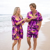 A man and a woman walking hand in hand on a sandy beach, wearing matching tropical print swim cover-ups in vibrant purple, yellow, and black.