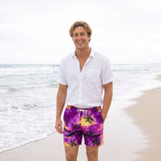 A man walking on a sandy beach wearing a white short-sleeve button-up shirt and purple tropical print swim trunks.
