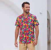 A man wearing a vibrant, multicolored tropical floral print short-sleeve button-down shirt and khaki pants, standing against a clean white stucco wall.