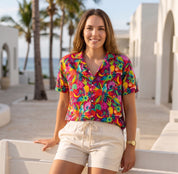 A woman stands outdoors in a sunny, Mediterranean-style setting wearing a vibrant, multicolor tropical floral print short-sleeve button-up shirt and cream-colored shorts.
