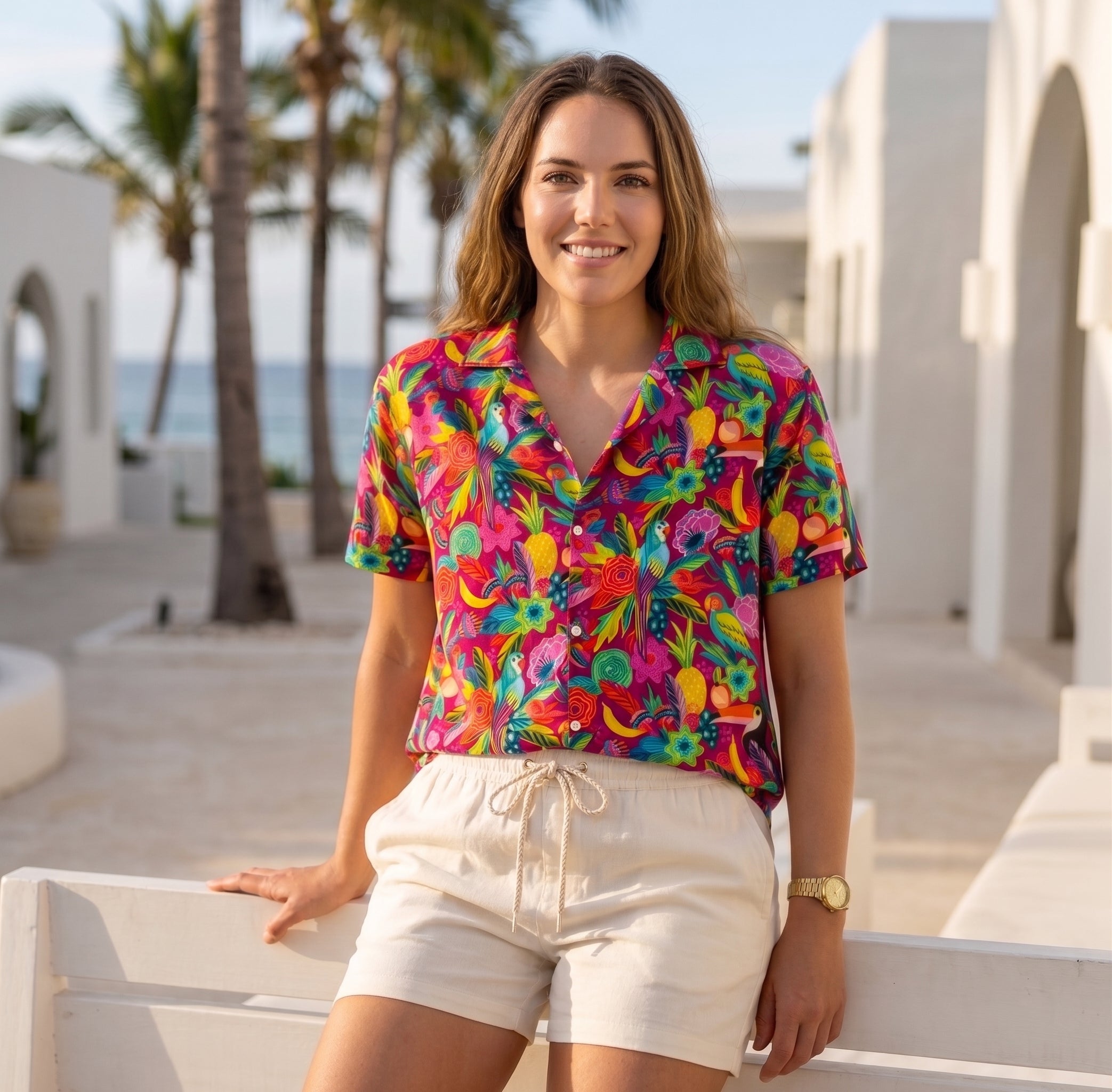 A woman stands outdoors in a sunny, Mediterranean-style setting wearing a vibrant, multicolor tropical floral print short-sleeve button-up shirt and cream-colored shorts.
