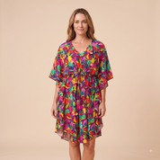 A woman modeling a short, V-neck dress with a colorful abstract floral print in shades of pink, purple, green, yellow, and blue, set against a solid beige studio background.