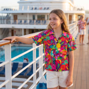 A young girl stands on a cruise ship deck wearing a vibrant, multicolored tropical floral short-sleeve button-up shirt and white shorts, with a pool and other passengers blurred in the background.
