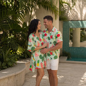 A man and a woman wearing matching tropical-print short-sleeve shirts and a matching short dress stand together in a sunlit outdoor patio setting with palm leaves and a tiled pool area in the background.