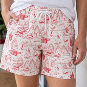 A woman stands on a wooden porch wearing a white sleeveless top and white shorts with a red and pink toile-style print. The background shows a white house exterior with holiday greenery.