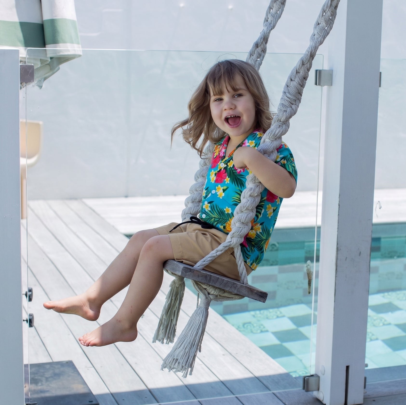 A young girl with brown hair sits smiling on a rope swing attached to a white patio pergola next to a swimming pool.