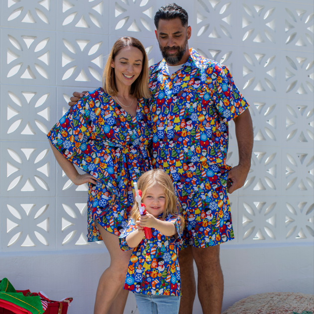 A family of three wearing matching colorful cartoon-print short-sleeve robes and shorts stands together in front of a white decorative wall. The set includes a baby/toddler, a woman, and a man, all in matching blue-based outfits with red, yellow, green, and orange character motifs.