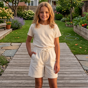 A young girl stands on a wooden boardwalk in a lush, well-maintained backyard garden, wearing a matching cream-colored short-sleeve t-shirt and shorts set.