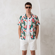 A man wearing a white short-sleeve button-up shirt with a colorful tropical floral print, white shorts, and white sneakers, standing against a textured light gray wall.
