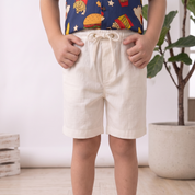 A child standing indoors wearing light cream drawstring shorts, a colorful patterned top, and cream slip-on shoes. The photo is cropped from the chest down, with a potted plant and wood floor in the background.
