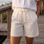 A woman stands on a wooden boardwalk by the beach wearing a white short-sleeve button-front shirt and white drawstring shorts. Palm trees, ocean, and a beach house are visible in the background.