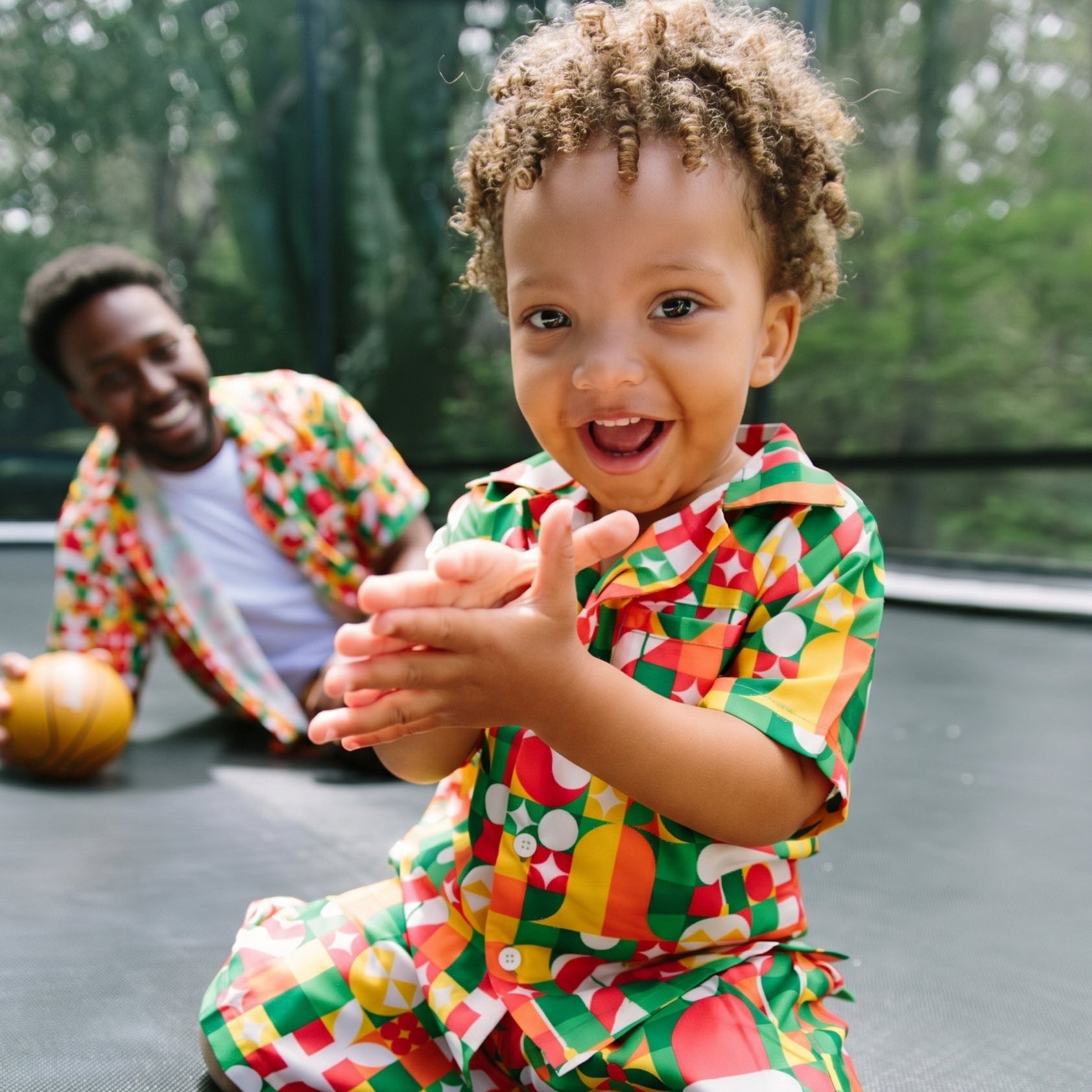 A toddler wearing a colorful geometric print matching shirt and pants sits on a trampoline, smiling and clapping. A man in a matching set is visible in the background.