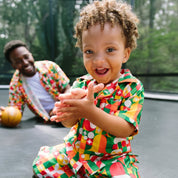A toddler wearing a colorful geometric print matching shirt and pants sits on a trampoline, smiling and clapping. A man in a matching set is visible in the background.