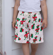 A young girl with blonde hair stands barefoot against a white wall and shutter, wearing a white t-shirt and cream shorts with a red and green frog pattern.