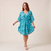 A woman modeling a turquoise blue holiday-themed kaftan dress with a Santa Claus and Christmas tree pattern, set against a solid beige studio background.