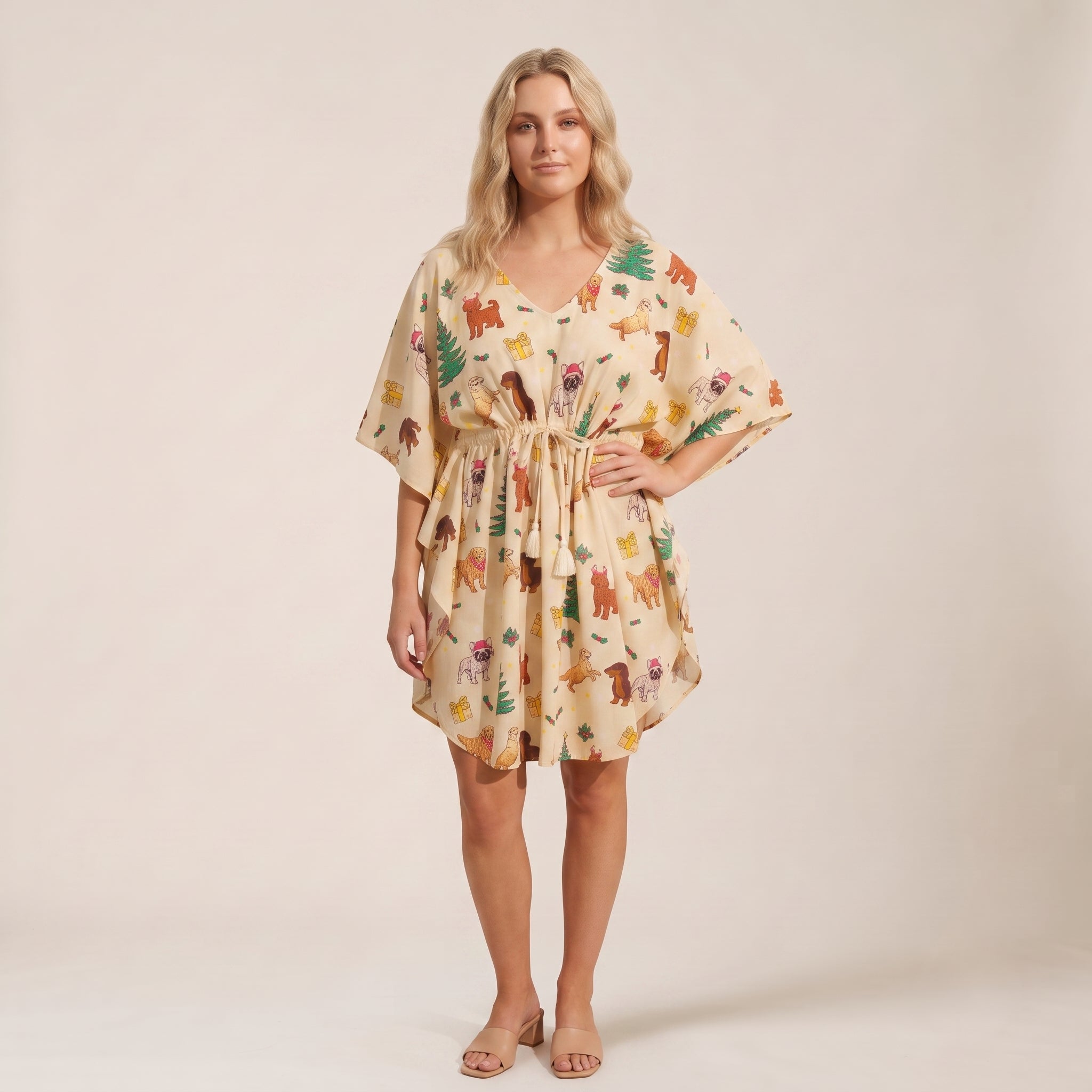 A woman modeling a short, loose V-neck dress with wide sleeves and a drawstring waist, covered in a colorful animal print on a cream background. She is standing against a plain beige studio backdrop.