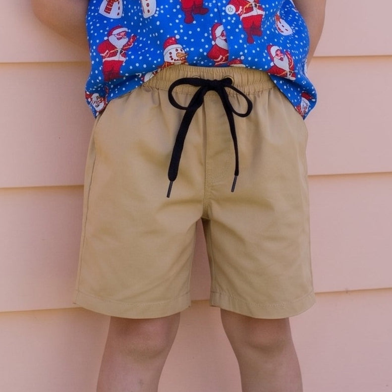 A young boy with blonde hair stands against a peach-colored house siding, wearing a blue short-sleeve button-up shirt with a Santa Claus pattern, tan shorts, and white sneakers.